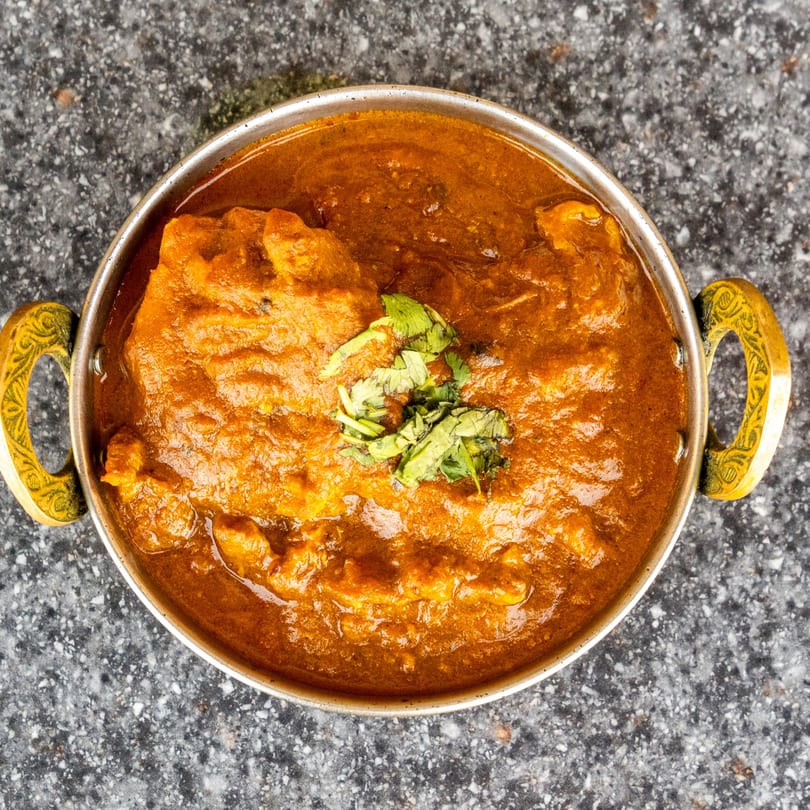 Top-down view of a metal bowl filled with rich orange curry, garnished with fresh green herbs.
